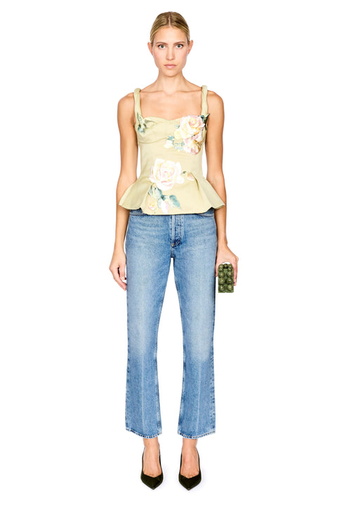 A model stands against a white background wearing a pale yellow-green floral camisole top, blue jeans, and dark green heels, while holding a small green clutch.