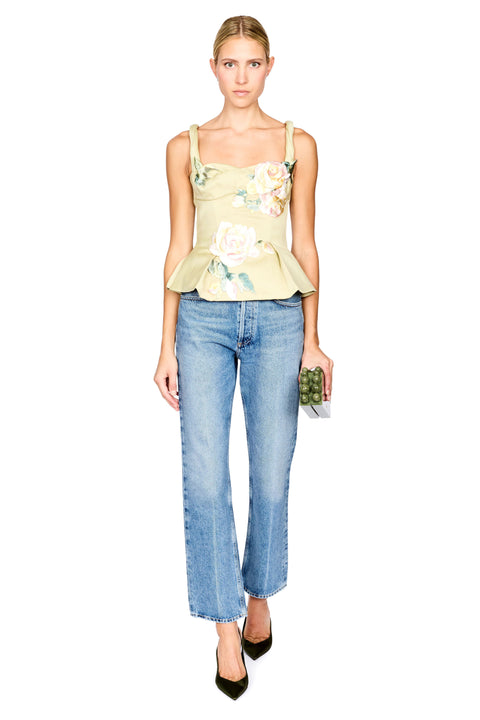 A model stands against a white background wearing a pale yellow-green floral camisole top, blue jeans, and dark green heels, while holding a small green clutch.