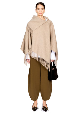 A model stands against a white background wearing a beige asymmetrical fringed wrap poncho over a white shirt, olive green harem pants, and black pointed flats, while holding a black handbag.