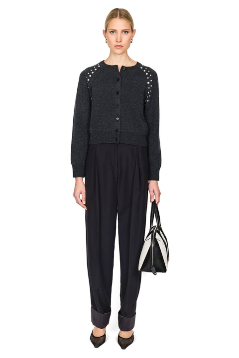 A model stands against a white studio background wearing a dark charcoal gray knit cardigan with silver stud embellishments on the shoulders, black pleated trousers, and holding a black and white handbag.