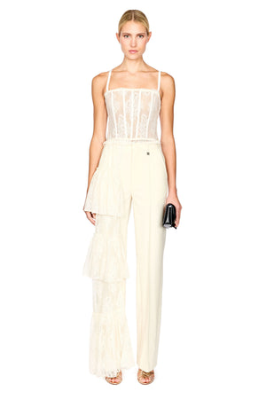 A model wearing a cream-colored two-piece outfit consisting of a sheer lace camisole top and matching high-waisted wide-leg trousers with a ruffled overlay, holding a small black clutch against a plain white background.