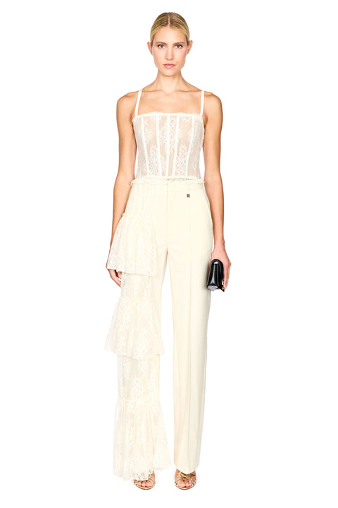 A model wearing a cream-colored two-piece outfit consisting of a sheer lace camisole top and matching high-waisted wide-leg trousers with a ruffled overlay, holding a small black clutch against a plain white background.