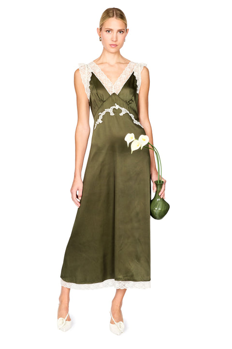 A woman models a sleeveless olive green satin slip dress with white lace trim at the V-neckline and hem, holding a small green clutch vase with white flowers against a plain white background.