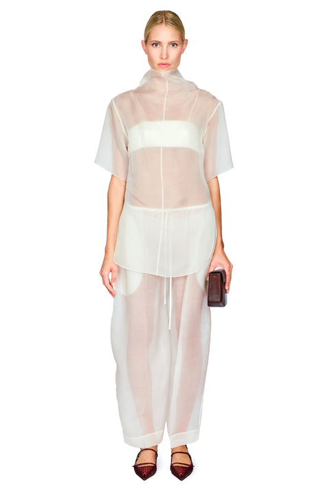 A model wearing a sheer white high-neck short-sleeve top with an attached bra insert and matching wide-leg sheer trousers, holding a small dark clutch against a plain white background