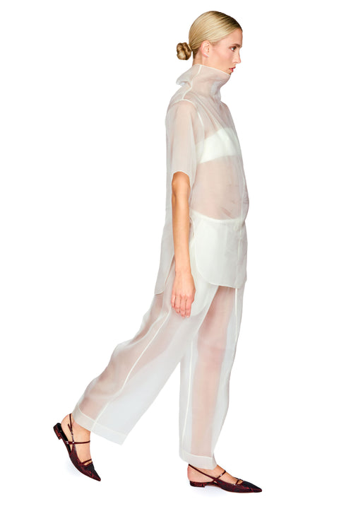 A model in profile wearing a sheer white high-neck tunic top and matching wide-leg sheer trousers, paired with dark brown pointed flats, against a solid white background.
