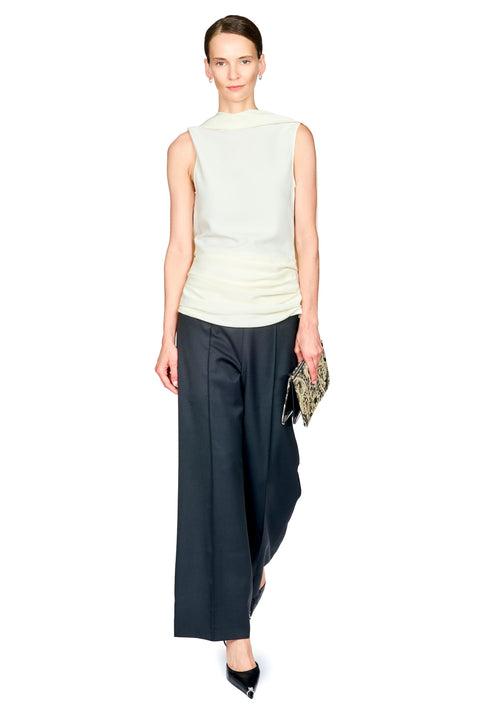 A model stands against a plain white background wearing a sleeveless white mock-neck top, dark black wide-leg trousers with white waist ruching, and black pointed heels, while holding a small patterned clutch.