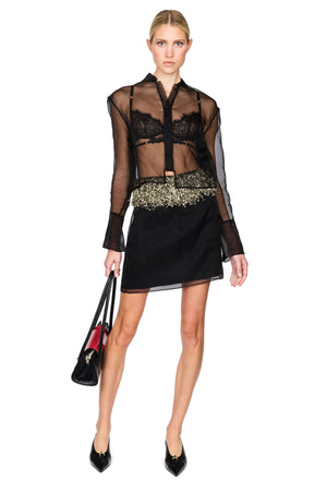 A model wearing a sheer black long-sleeve blouse with a black lace bralette overlay and a black satin mini skirt featuring a gold patterned waistband, holding a black handbag against a solid white background.