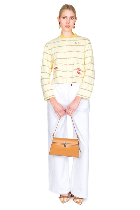 Woman wearing a yellow striped sweater with red fish on the pockets and white pants on a white background clutching bag with both hands.