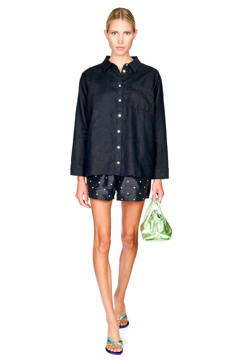 A model walks against a white background wearing a black long-sleeve button-up shirt, black shorts with white star patterns, and holding a green patterned plastic bag.