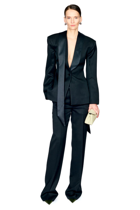 A model wearing a black satin-look tailored suit with dramatic shoulder peaks, a deep V-neckline, and straight-leg trousers, holding a small light beige clutch against a plain white background.