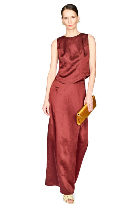 A woman modeling a sleeveless rust-red sleeveless top and pants set holding a gold clutch against a plain white background.