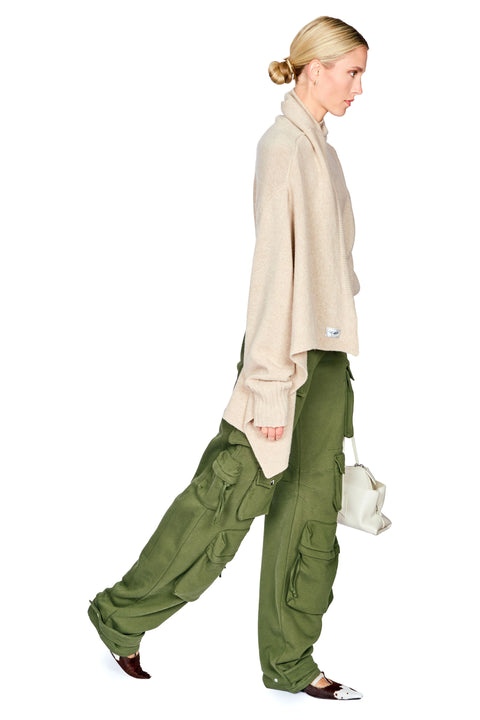 A woman in profile wearing an oversized beige knit sweater, olive green cargo pants, and dark pointed cow flats, holding a small white handbag against a plain white background.