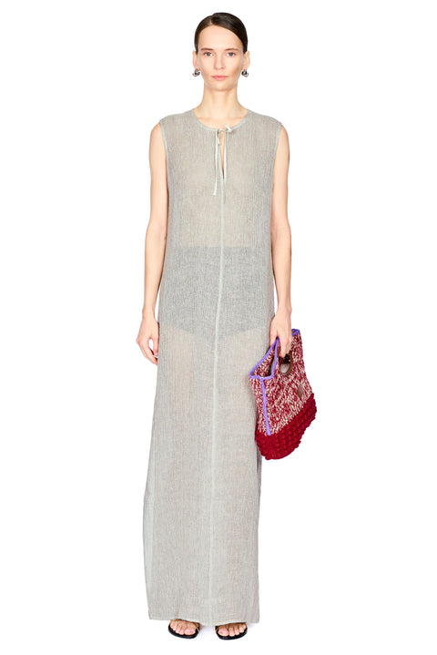 A model wearing a sleeveless, light gray linen maxi dress with a tie-neck detail, holding a red and purple patterned woven clutch bag against a plain white background.