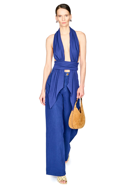 A model wearing a royal blue halter-neck top with wide-leg pants, holding a tan leather handbag against a plain white background.