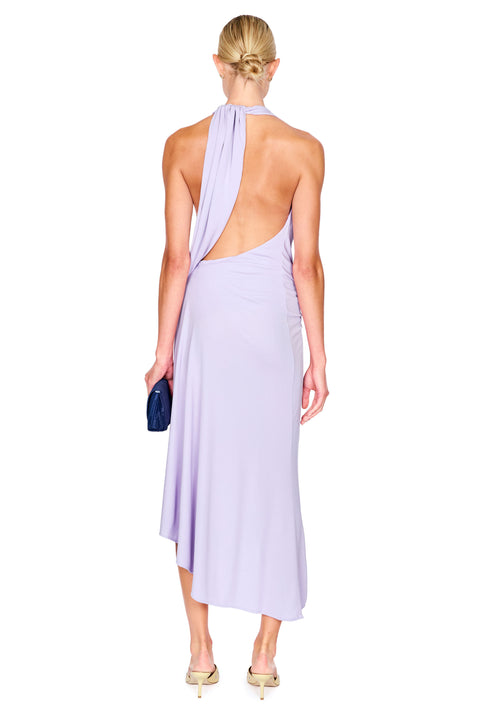 A woman shown from the back wearing a lavender halter-neck midi dress with a dramatic open back and draped neckline detail. She is holding a small dark blue clutch and wearing beige heels, set against a plain white background.