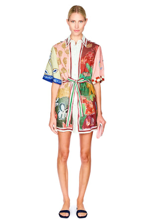 A model wearing a short-sleeved, collared mini dress with a multicolor abstract print, featuring a tie waist belt and striped trim, paired with black slide sandals against a plain white background.
