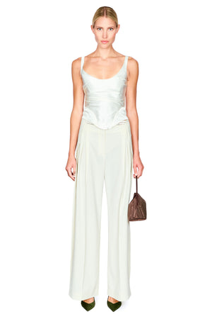 A model stands against a white background wearing a white satin camisole top and matching wide-leg trousers, paired with olive green pointed shoes and holding a brown leather handbag.