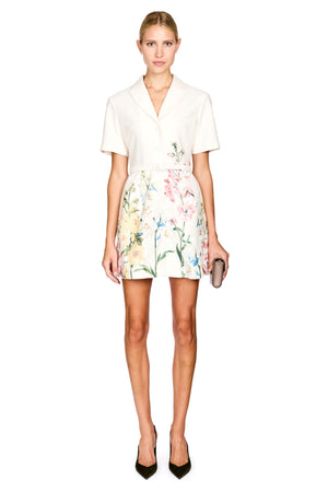 A woman modeling a short-sleeve white collared top paired with a high-waisted white mini skirt featuring a pastel floral print, holding a small metallic clutch and wearing black pointed-toe heels against a plain white background.