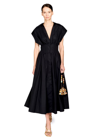 A model wearing a black midi dress with a deep V-neckline, draped cap sleeves, and a gathered waist, styled with gold sandals and a gold beaded handbag against a plain white background.