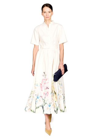 A model wearing a cream-colored short-sleeve midi dress with a floral watercolor print at the hem, holding a dark navy clutch, set against a plain white background.