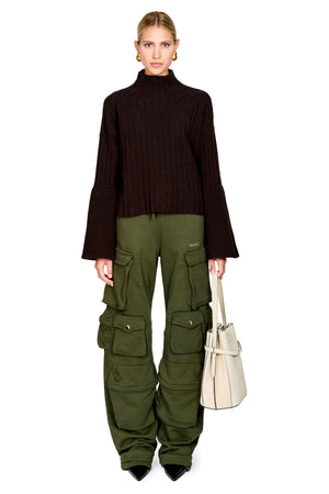 A model stands against a white background wearing a dark brown ribbed turtleneck sweater, olive green cargo pants, and holding a cream-colored tote bag.