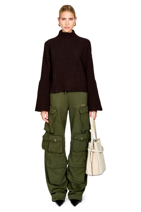 A model stands against a white background wearing a dark brown ribbed turtleneck sweater, olive green cargo pants, and holding a cream-colored tote bag.