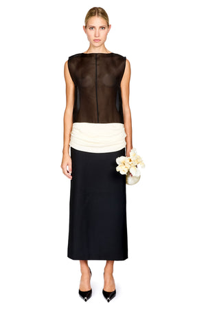 A woman standing in front of a white background wearing a black and white colorblocked long dress with a sheer top and white ruched waistband