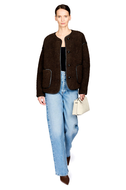A model wearing a dark brown bouclé button-front jacket with contrast piping, light-wash blue jeans, a black top, and brown pointed shoes, holding a small cream handbag against a plain white background.
