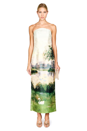 A woman modeling a strapless maxi dress with a scenic landscape print featuring a lake, trees, and a white swan, paired with nude high heels and a small clutch, against a plain white background.