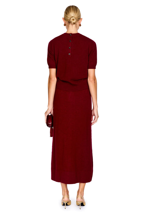A woman viewed from the back wearing a short-sleeve, high-neck, deep burgundy knit top and matching midi skirt, holding a small matching clutch against a plain white background.
