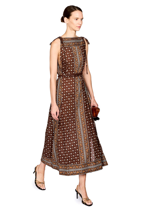 A woman modeling a sleeveless brown midi dress with white polka dots and gold patterned borders, paired with gold heeled sandals and holding a small brown clutch, set against a plain white background.