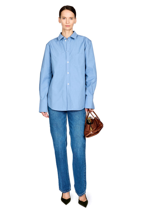 A model stands against a white background wearing a light blue long-sleeve button-up shirt, blue jeans, and black pointed heels, while holding a brown leather handbag.