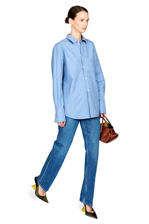 A woman modeling a light blue long-sleeve button-up shirt, blue jeans, and olive green pointed-toe heels, holding a brown woven handbag against a plain white background.