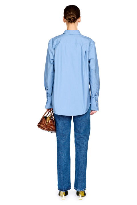 A model shown from the back wearing a light blue long-sleeve button-up shirt and blue jeans, holding a brown handbag against a plain white background.
