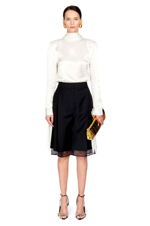 A model stands against a white studio background wearing a white satin mock-neck long-sleeve top, a black pleated midi skirt with sheer side panels, black strappy high-heeled sandals, and holding a gold clutch.