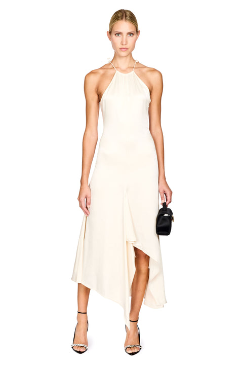 A woman modeling a cream-colored halter-neck midi dress with an asymmetrical draped hem, paired with black high-heeled sandals and a small black handbag, set against a plain white background.