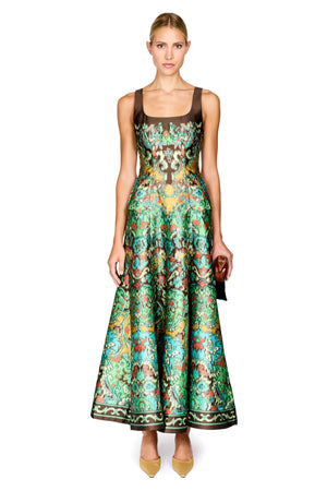 A model wearing a sleeveless, floor-length A-line dress with a brown square neckline and a vibrant green, gold, and red patterned skirt, paired with tan pointed-toe heels and holding a small black clutch, set against a plain white background.