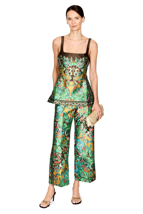 A model stands against a white background wearing a matching two-piece set consisting of a sleeveless peplum top and wide-leg pants, both featuring a vibrant green, blue, and orange floral pattern with a dark brown mesh underlayer. She is holding a small beige clutch and wearing gold sandals.
