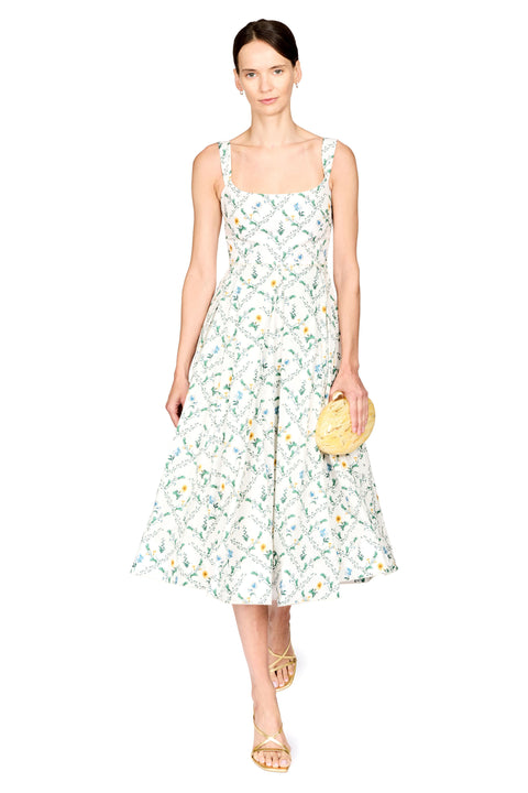 A woman modeling a sleeveless midi dress with a white base and a green and yellow botanical pattern, holding a round yellow clutch against a plain white background.