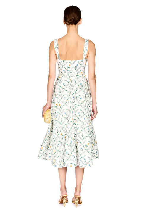 A woman shown from the back wearing a sleeveless midi dress with a white base and an allover green and yellow botanical pattern. She is holding a small gold clutch and wearing gold heels against a plain white background.