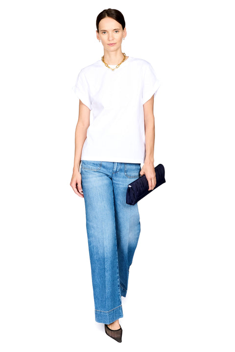 A woman modeling a white short-sleeve t-shirt and wide-leg blue jeans, holding a dark navy clutch purse against a solid white background.