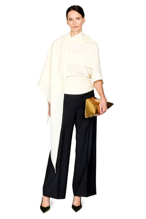A model wearing a cream draped wrap top, black wide-leg trousers, and black pointed heels, holding a gold textured clutch against a plain white background.