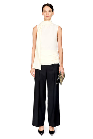 A model stands against a white background wearing a sleeveless cream mock-neck top with a draped collar, black wide-leg trousers, and black pointed heels, while holding a small patterned clutch.