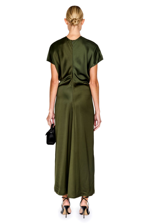 A woman viewed from the back wearing a satin-like olive green maxi dress with short sleeves and a gathered back detail. She is standing against a plain white background and holding a small black handbag.