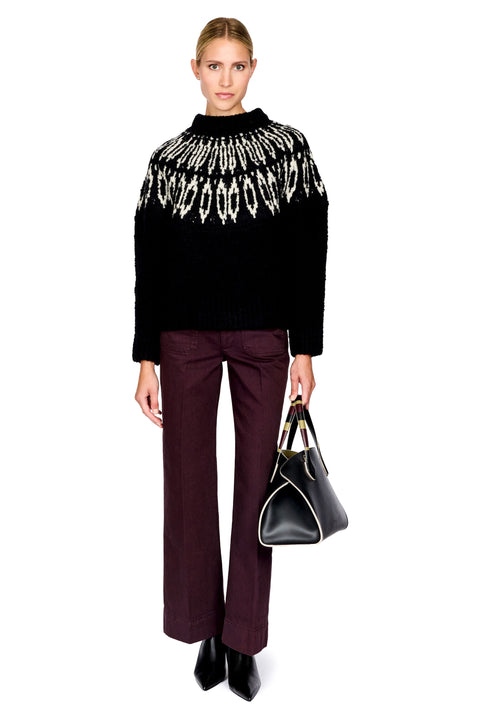 A model stands against a white studio background wearing a black knit sweater with a white geometric pattern across the upper chest, dark plum wide-leg trousers, black pointed shoes, and carrying a black handbag with gold trim.