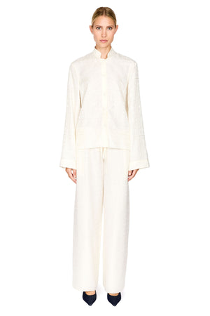 A model stands against a solid white background wearing a matching cream-colored two-piece set consisting of a long-sleeve cropped jacket with a mandarin collar and wide-leg trousers.