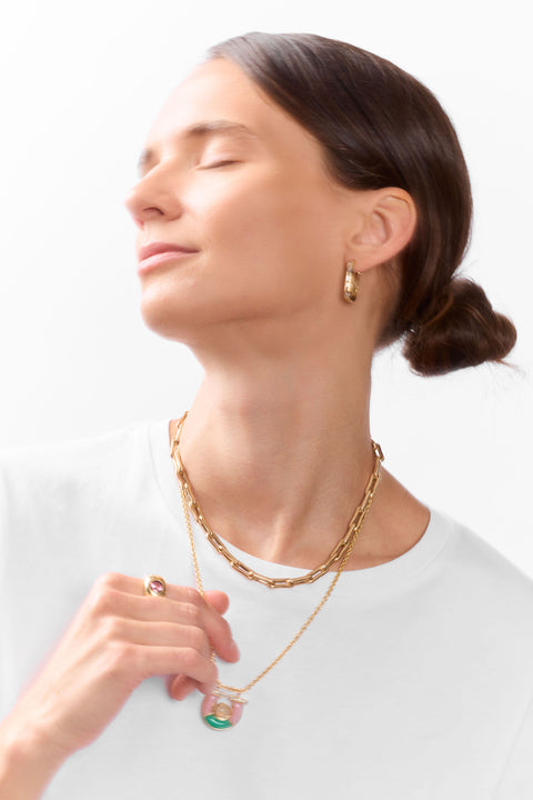 A woman with her eyes closed wears a gold chain necklace with a colorful horseshoe pendant, a matching gold ring, and a small gold hoop earring against a clean white background.