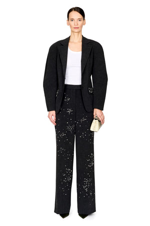 A model stands against a white studio background wearing a black textured blazer, matching wide-leg trousers with silver bead embellishments, a white tank top, and holding a small beige clutch.