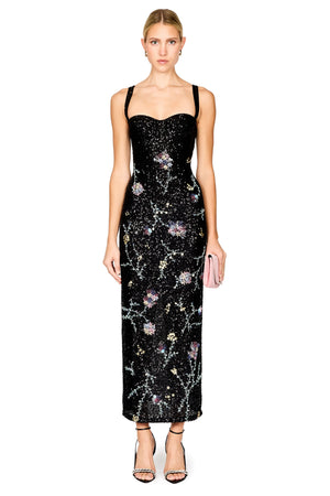 A model wearing a floor-length black sequined gown with floral embroidery and thin shoulder straps, paired with black strappy heels and holding a small pink clutch, against a plain white background.