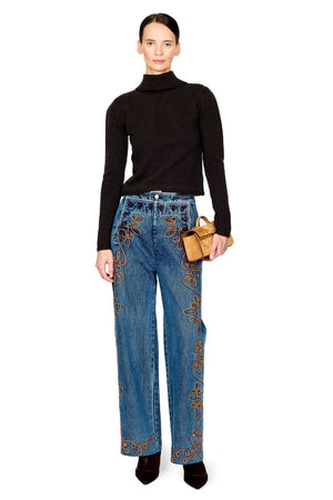 A model stands against a white background wearing a brown turtleneck sweater, blue wide-leg jeans with gold embroidery, and holding a small brown leather bag.